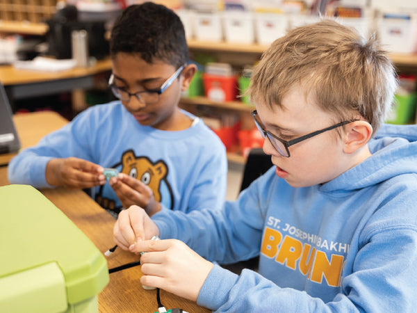 Two students wearing glasses assembling a green electronic component at a wooden desk | Forward Education
