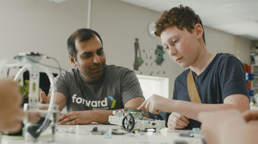 Instructor guiding student assembling white robotics vehicle with wheels and circuit board components | Forward Education